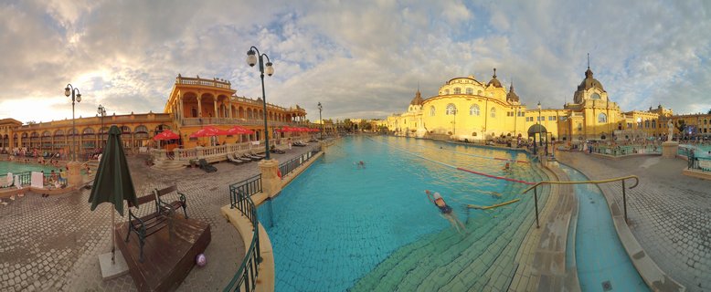 Panoramic view of outdoor thermal pool with swimmers at Széchenyi Bath, surrounded by yellow historic buildings with domes under cloudy sky at sunset