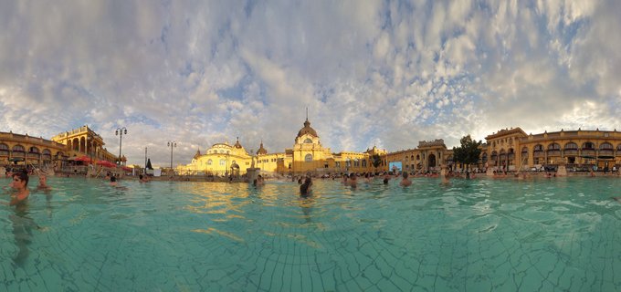 Wide view of people swimming in turquoise thermal pool with ornate yellow Neo-Baroque Széchenyi Bath building and cloudy sky in Budapest