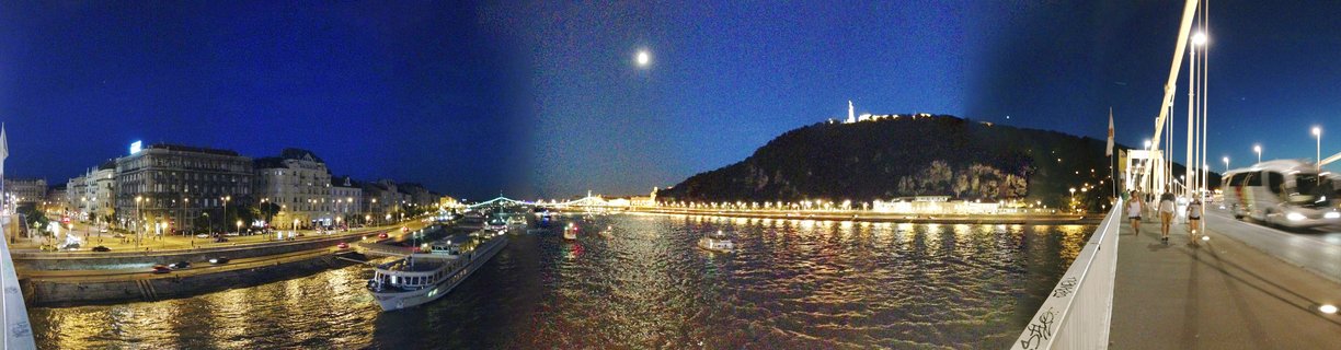 Panoramic night photograph of Budapest showing the Danube River with illuminated buildings, Gellért Hill with the Liberty Statue on top, Elisabeth Bridge with people walking, boats on the water, and a full moon in the dark blue sky
