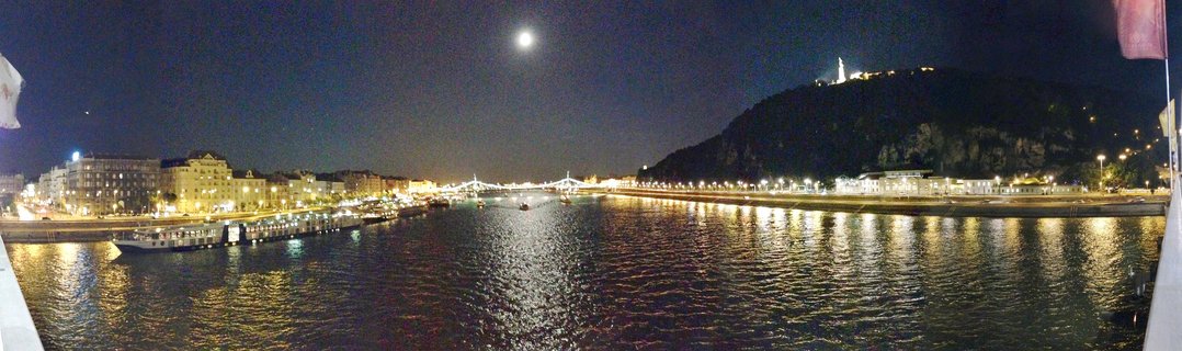 Panoramic night photograph of Budapest showing the Danube River with illuminated buildings, Liberty Bridge, Gellért Hill with the Liberty Statue at its summit, and a full moon in the dark sky with light reflections on the water