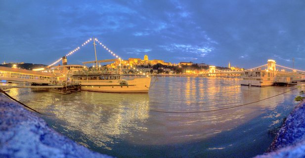 Panoramic view of illuminated Buda Castle and Chain Bridge over the Danube River at twilight in Budapest, with boats docked along the waterfront and city lights reflecting on the water