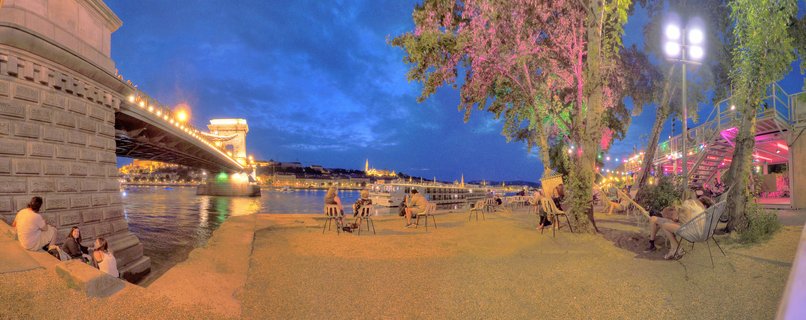 Panoramic night view of Budapest's Chain Bridge over the Danube River with people sitting at an outdoor riverside bar with colored tree lighting