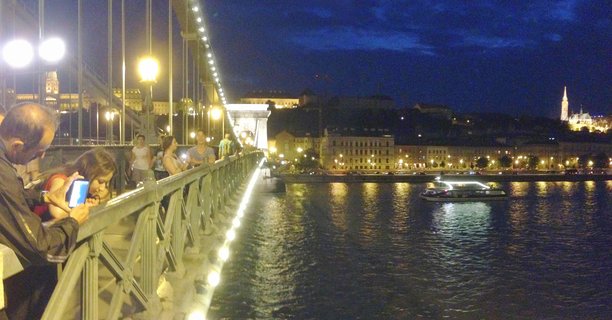 Night view of Budapest's Chain Bridge with people on the walkway, illuminated Buda Castle on the hill, Matthias Church spire, and a lit tour boat on the Danube River under a deep blue twilight sky.