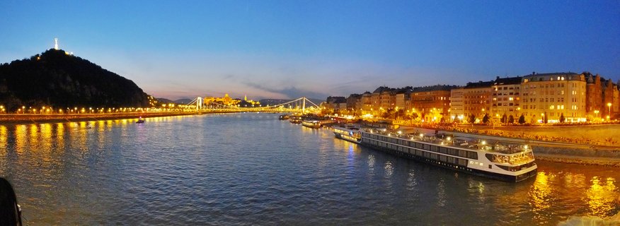 Panoramic cityscape of Budapest at twilight showing the Danube River, illuminated Chain Bridge, Gellért Hill with Liberty Statue, and river cruise ships with city lights reflecting on water