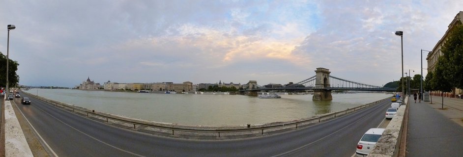 Wide panoramic photograph of Budapest showing the Danube River with the Chain Bridge spanning across, Parliament Building visible on the left bank, cars on the riverside road, and cloudy sky with warm sunset tones