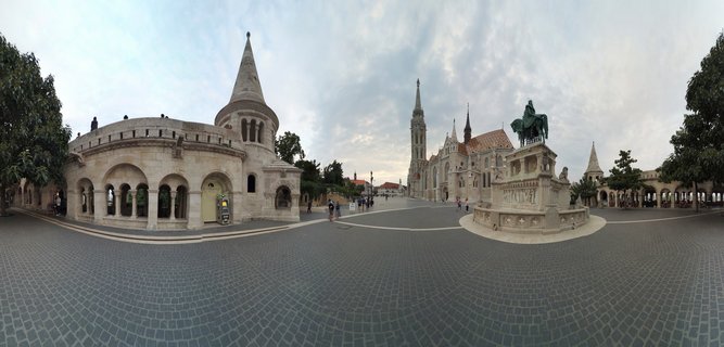 Panoramic view of Fisherman's Bastion's white stone towers and Matthias Church's Gothic spire at Castle Hill in Budapest, with cobblestone plaza and Saint Stephen equestrian statue under cloudy sky