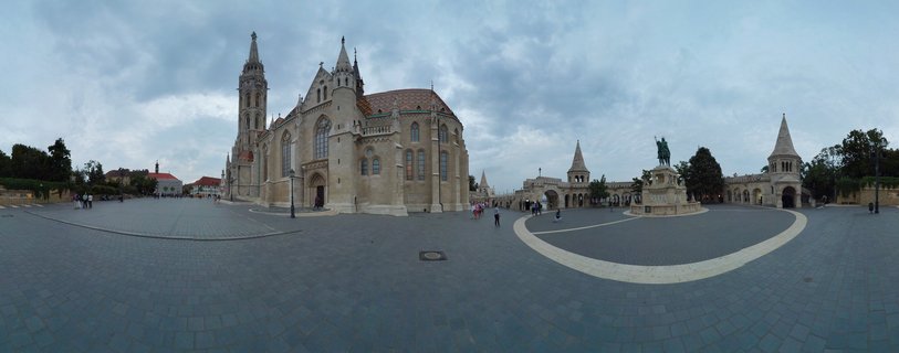 Wide view of Matthias Church with its tall Gothic spire and Fisherman's Bastion with conical towers on Castle Hill in Budapest, with tourists walking on the paved plaza under cloudy sky