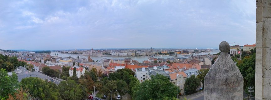 Wide panoramic cityscape of Budapest showing the Danube River, Hungarian Parliament Building, red-roofed buildings, and green trees under cloudy sky with stone architectural element in foreground