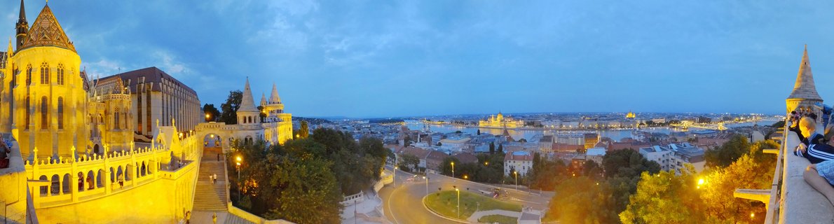 Panoramic view of illuminated Fisherman's Bastion towers at twilight overlooking Budapest cityscape with Hungarian Parliament Building visible across the Danube River