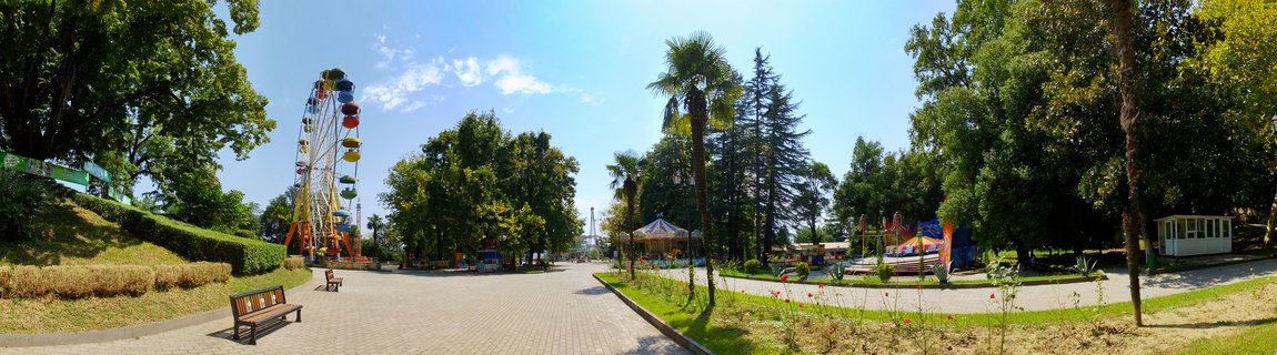 A colorful Ferris wheel at an amusement park in Kutaisi, Georgia, with paved walkways, benches, palm trees, and other carnival rides visible under a clear blue sky
