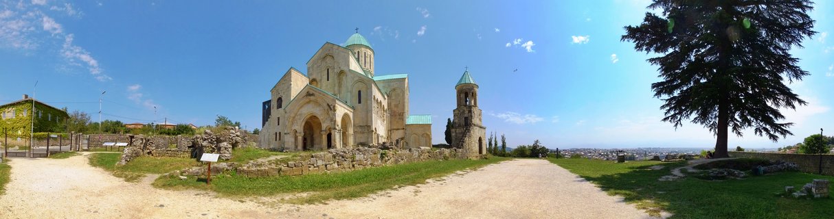 Panoramic view of Bagrati Cathedral, a medieval stone church with green domes and a bell tower, surrounded by ruins and pathways under a blue sky in Kutaisi, Georgia
