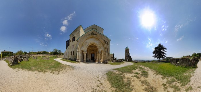 Medieval stone cathedral with arched entrance and bell tower surrounded by ruins and gravel pathways under bright blue sky in Kutaisi, Georgia