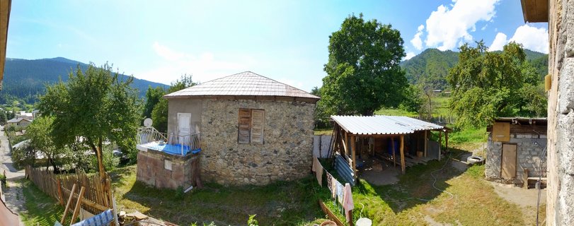 Stone houses with metal roofs in a mountain village with green trees, blue sky, and mountains in the background