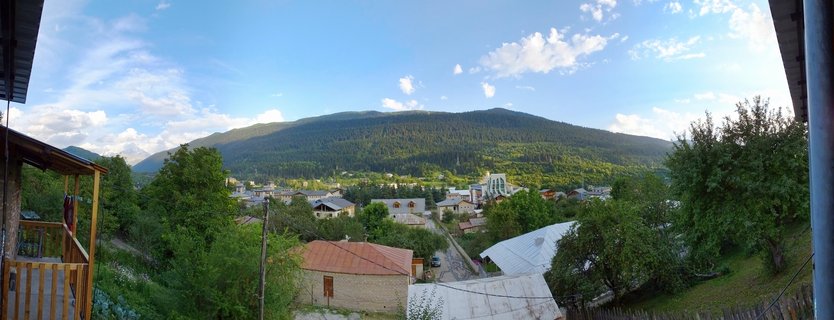 Panoramic landscape showing the town of Mestia in Georgia's Svaneti region, with residential buildings in the foreground, forested mountains rising in the background, and blue sky with white clouds above
