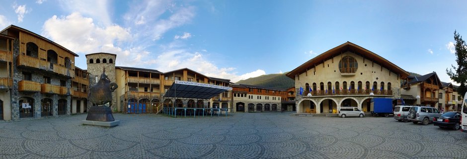 Panoramic view of a cobblestone town square in Mestia, Georgia, surrounded by medieval Svan tower houses with wooden balconies, featuring a bronze statue and mountains in the background under a blue sky with clouds