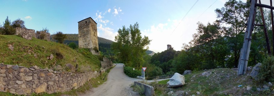 Medieval stone Svan tower on grassy hillside with dirt road, stone wall, trees, and mountain forest background in Mestia, Georgia