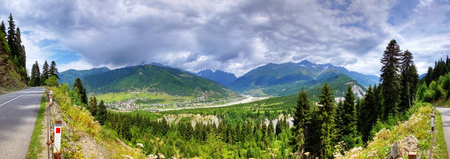 Wide panoramic landscape showing a green mountain valley with a town, forested hillsides with pine trees, a winding road, and distant mountain ranges under a cloudy sky in Mestia, Georgia
