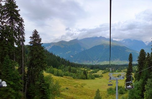 View from a chairlift showing empty lift chairs suspended on cables above green mountain meadows with pine trees and layered blue mountain ranges under cloudy sky