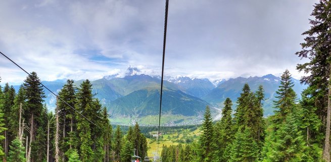 View from cable car showing green pine forest, mountain valley with town, and snow-capped Caucasus mountains under cloudy sky