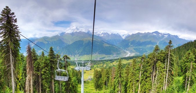 Chairlift cables and empty seats rise through green pine forest toward snow-capped mountain range with cloudy sky above Mestia valley in Georgia