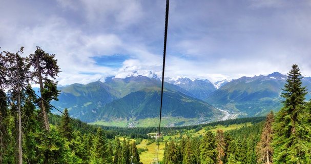 Panoramic mountain landscape showing green forested slopes, a valley with scattered buildings, and snow-capped Caucasus peaks under partly cloudy sky, viewed from cable car with visible cable