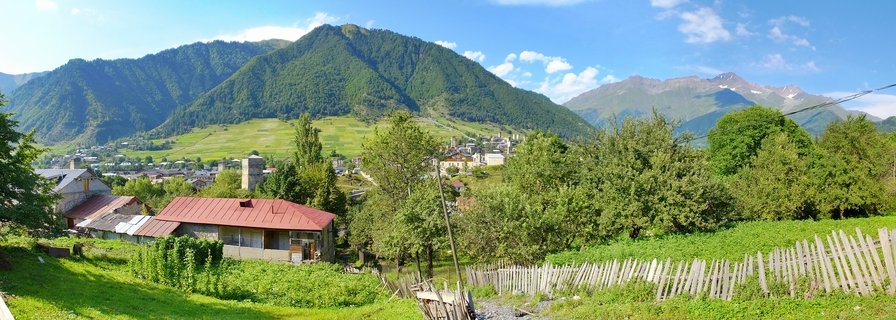 Panoramic view of Mestia village showing traditional stone tower houses scattered across green mountain valley with forested slopes and distant peaks under blue sky