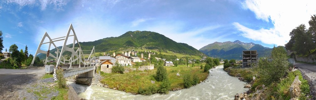 Panoramic view of Mestia, Georgia showing medieval stone tower houses, a white truss bridge, rushing river, and green Caucasus mountains under blue sky