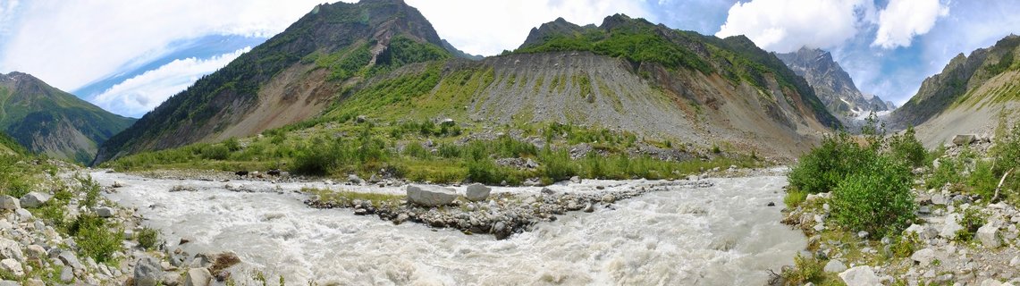 Mountain landscape showing a fast-flowing river with white water rapids in the foreground, rocky banks with green shrubs, steep mountain slopes with exposed rock and vegetation, and distant snow-capped peaks under a partly cloudy sky