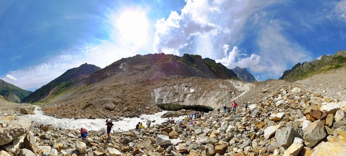Panoramic view of hikers on rocky glacier moraine with ice cave formation, mountain peaks, and meltwater stream under blue sky in Mestia, Georgia