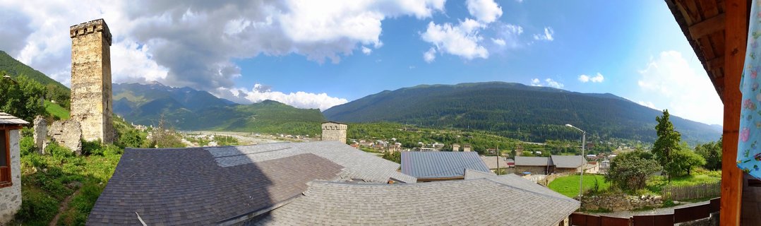 Stone medieval defense towers overlooking village rooftops with forested mountains and cloudy blue sky in Mestia, Georgia