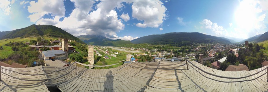 Panoramic landscape showing medieval stone towers in a green mountain valley with forested slopes and blue sky with white clouds in Mestia, Georgia