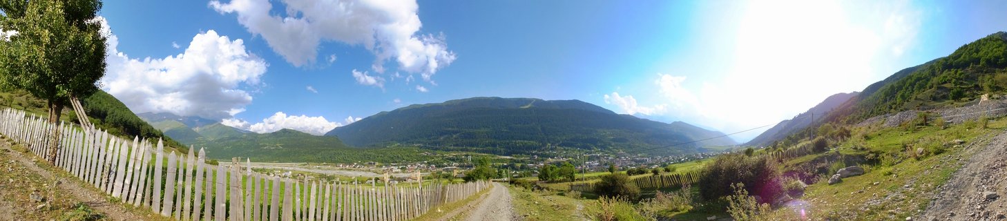 Wide mountain valley landscape showing white picket fence, dirt road, green fields, village, and forest-covered mountains under blue sky with clouds in Mestia, Georgia