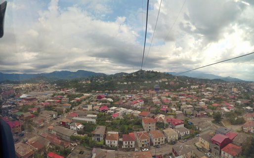 Aerial panoramic photograph showing Batumi city from a cable car with colorful residential rooftops, cable lines, green hills, and mountains under a cloudy sky