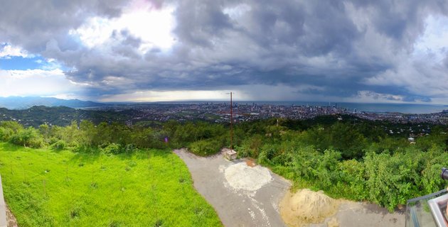 Panoramic landscape showing Batumi city and Black Sea coastline from an elevated green hillside viewpoint under dramatic cloudy sky with sun breaking through