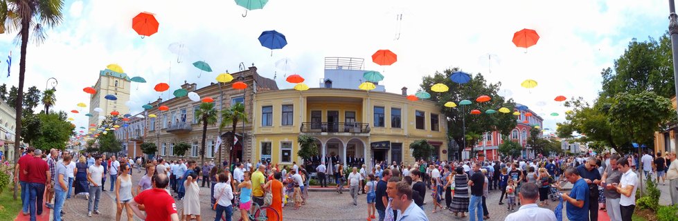 Wide street view showing crowds of people walking beneath colorful umbrellas suspended in the air above historic yellow and beige buildings in Batumi Georgia