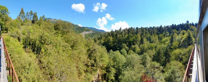 Panoramic view of green forest-covered mountains in Borjomi, Georgia with blue sky and white clouds, photographed from a bridge with red railings visible on both sides