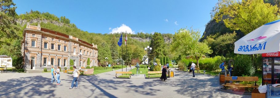 Wide panoramic view of a public square in Borjomi, Georgia, showing a historic two-story brick building, people walking on paved ground, wooden benches, flags, green trees, and forested mountains under a clear blue sky