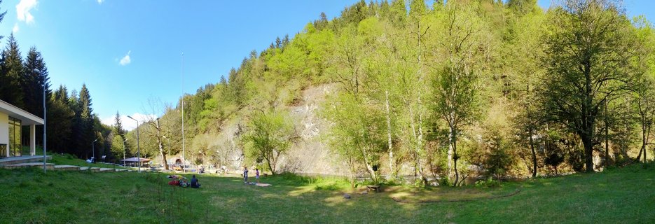 Wide landscape showing a grassy park area with people sitting on the ground, scattered green trees, a forest-covered hillside with exposed rock face, and a modern white building on the left under a blue sky