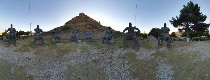 Panoramic view of bronze headless seated statues in foreground with historic fortress ruins on hilltop in background under clear evening sky