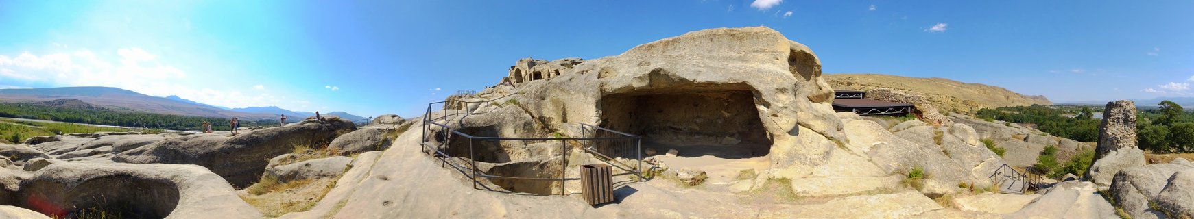 Panoramic view of Uplistsikhe ancient cave town showing rock-cut structures carved into sandstone cliffs with metal railings, visitors walking on pathways, arid landscape, distant mountains and blue sky with clouds