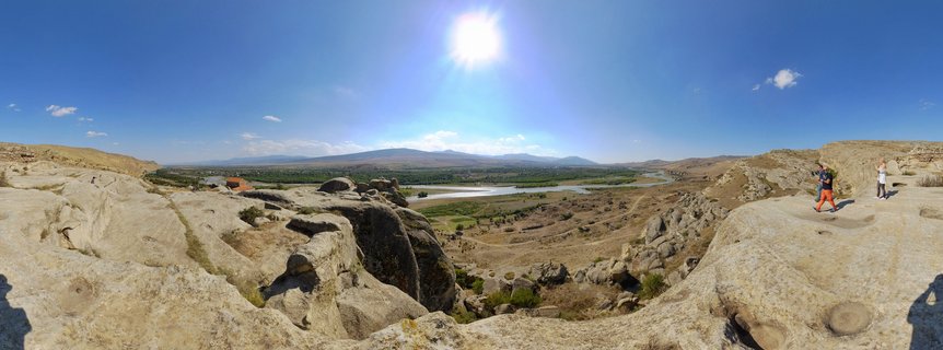 Wide panoramic photograph showing ancient rock-cut cave structures of Uplistsikhe in Georgia, with tourists walking on stone formations, a river valley below, and mountains in the distance under bright blue sky