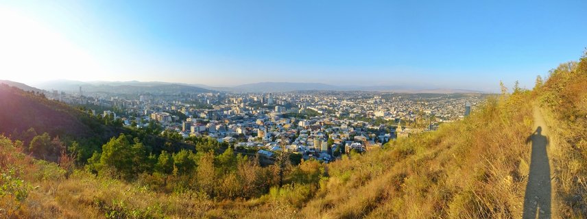 Wide panoramic photograph of Tbilisi city viewed from a grassy hillside, with urban buildings filling the valley, mountains in the distance, and clear blue sky above