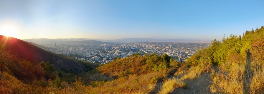 Panoramic cityscape of Tbilisi Georgia viewed from hillside during sunset with urban buildings in valley and mountains in background
