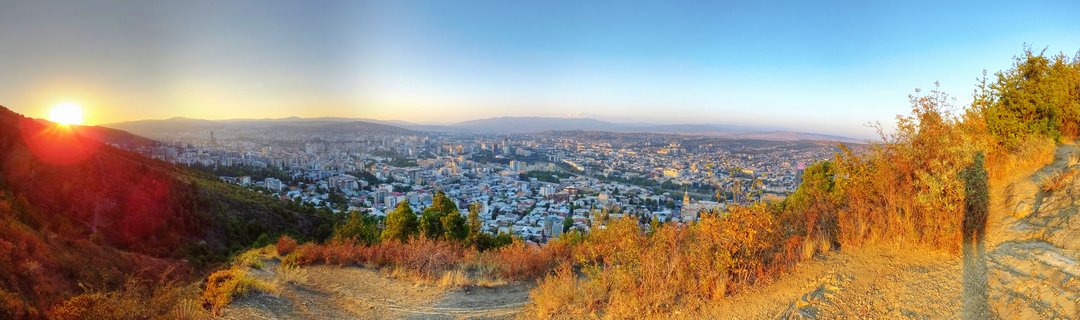 Panoramic cityscape of Tbilisi, Georgia at sunset showing dense urban buildings in the midground, dry golden grass and vegetation in the foreground, distant mountains on the horizon, and a clear blue sky with warm golden light from the setting sun on the left