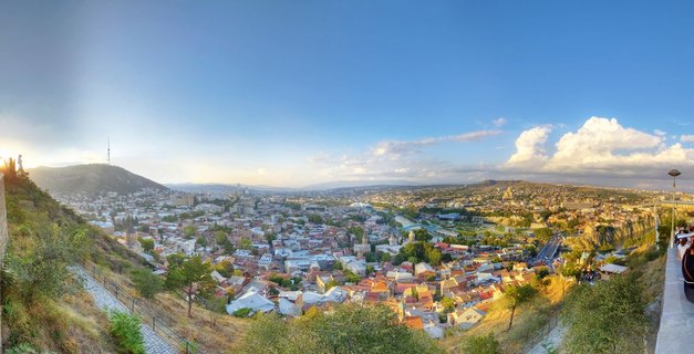 Wide panoramic photograph of Tbilisi cityscape showing dense urban area with red-tiled roofs, river valley, surrounding hills, and TV tower on mountain peak under blue sky with scattered clouds during golden hour