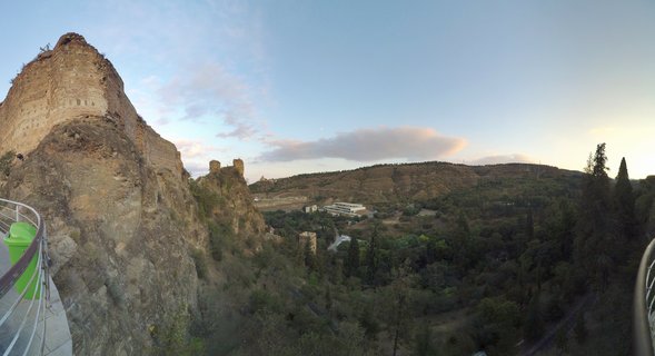 Panoramic view of ancient stone fortress ruins on a rocky cliff overlooking a green valley with trees and hills in Tbilisi, Georgia during sunset