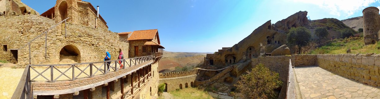 Panoramic view of ancient stone monastery buildings carved into rocky hillsides in Signagi, Georgia, with two people walking on a wooden walkway under blue sky