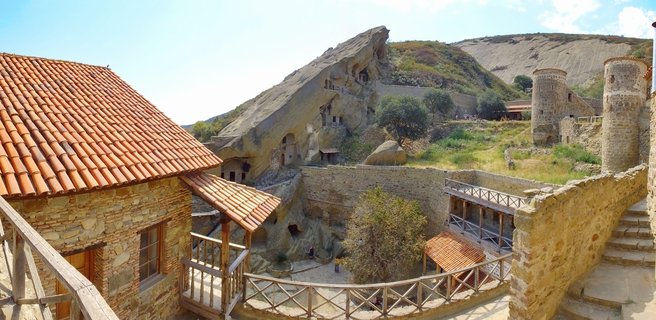 Stone monastery buildings with red tile roofs and cylindrical towers surrounding a courtyard with cave dwellings carved into a rocky hillside under blue sky