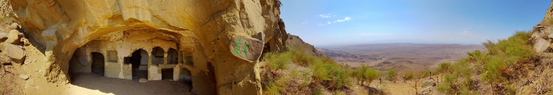 Panoramic view of ancient cave dwellings carved into a rocky cliff face with arched openings, green shrubs on hillside, and distant valley under blue sky in Georgia