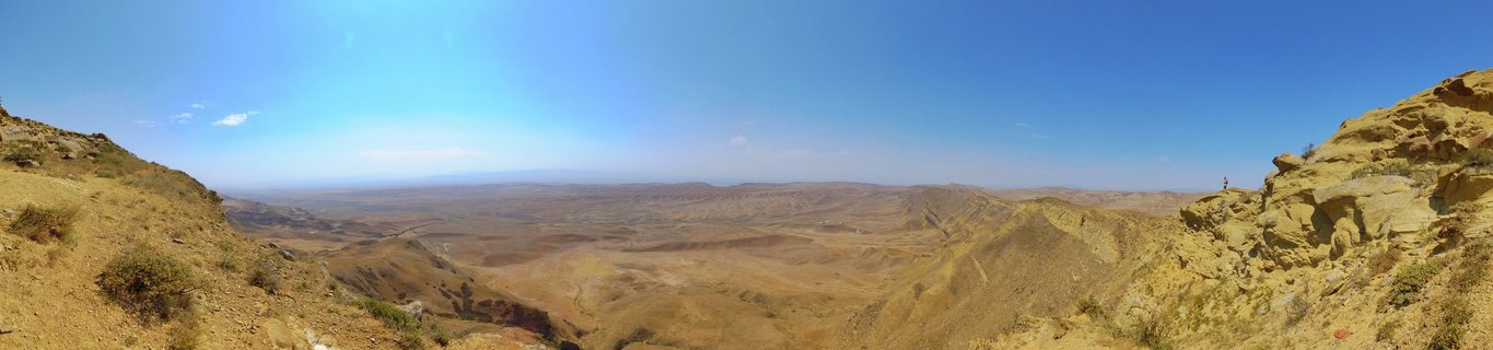Panoramic landscape showing a vast arid valley with golden-brown rolling hills, rocky outcrops in the foreground, and clear blue sky above the Alazani Valley near Signagi, Georgia